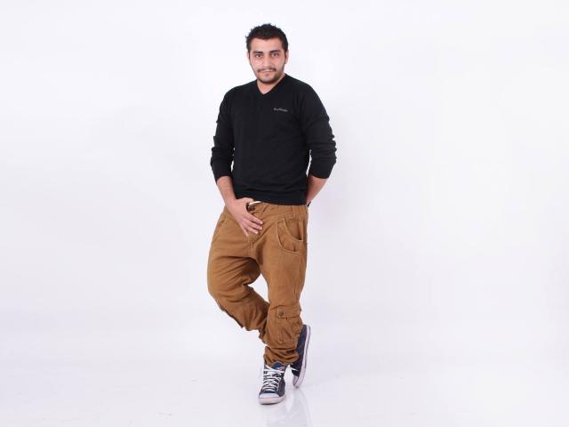 BrownHanson è in piedi con una gamba incrociata, indossa una maglietta nera a maniche lunghe, pantaloni cargo color sabbia e scarpe da ginnastica blu. Sta sorridendo, ha i capelli corti e scuri e una mano sulla parte inferiore del corpo..