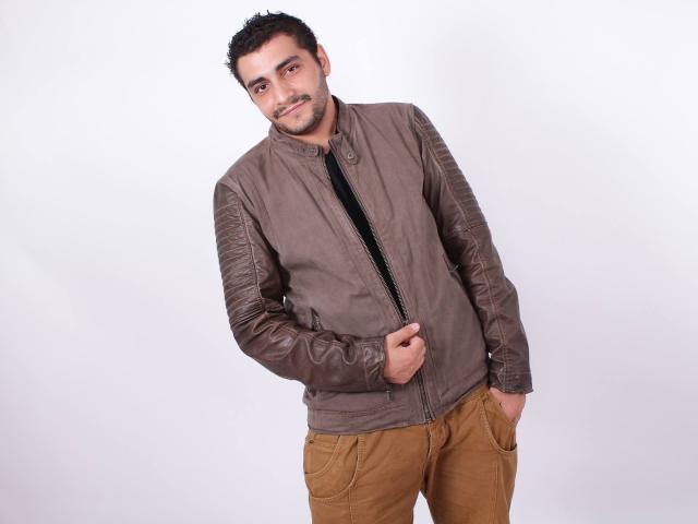BrownHanson, con capelli e barba corti e scuri, indossa una giacca di pelle marrone con maniche texturizzate e pantaloni color sabbia, in piedi e sorridente, con una mano in tasca, su sfondo bianco..