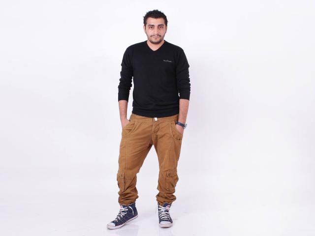 BrownHanson se tient debout, les mains dans ses pantalons de cargo beige, portant une chemise noire à manches longues et des baskets bleues, sur un fond blanc. Il a une courte barbe et des cheveux foncés..