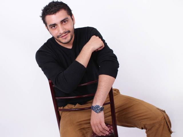 Un uomo di colore, con capelli neri ricci, barba e pelle scura, indossa una camicia nera a maniche lunghe e pantaloni beige, è appoggiato a una sedia rossa, sorride e indossa un orologio nero..