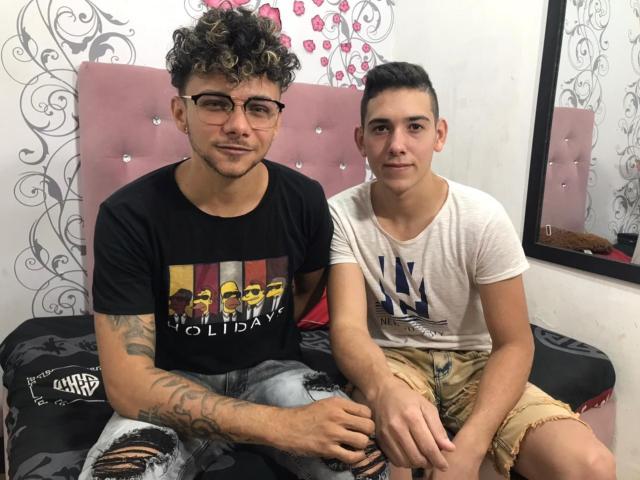 JorgeYCamiloHot with curly hair, glasses, and tattoos sits next to JorgeYCamiloHot with short hair, both wearing t-shirts and ripped jeans, on a pink bed.