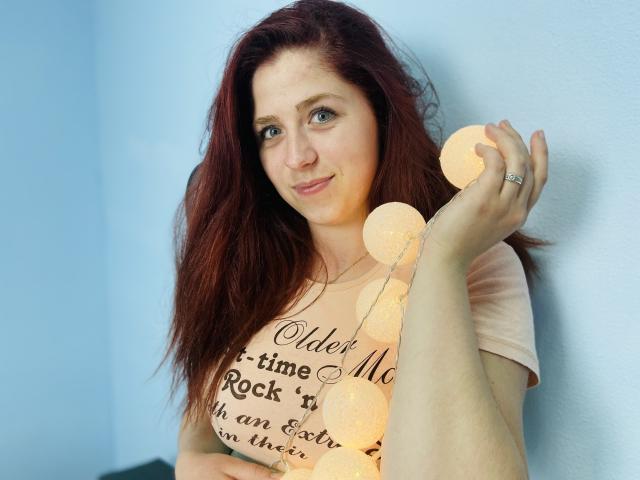 JessikaDaniels with red hair, holding warm string lights against a blue wall, wearing a beige shirt, smiling slightly, showing a ring on her right hand.