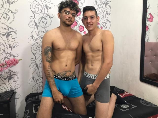 JorgeYCamiloHot with curly hair and glasses, wearing blue Calvin Klein underwear, has a tattoo on his right arm. JorgeYCamiloHot with short hair, wearing gray Calvin Klein underwear, smiles.