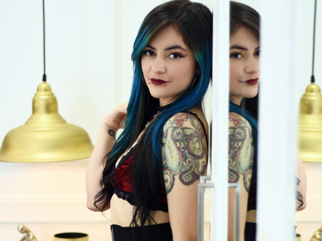 Candice Valo, com cabelo azul-preto, sutiã vermelho-preto, braço tatuado, batom escuro, olhando para o espelho, mão tocando o cabelo, luminária dourada ao fundo, reflexo mostrando a mesma expressão..