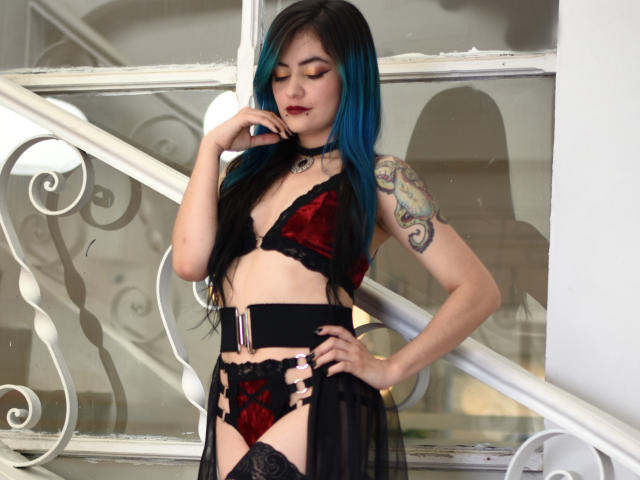 Candice Valo con reflejos azules, lencería de encaje negra y roja, collar negro, tatuaje en el brazo derecho, de pie junto a una escalera blanca, con la mano cerca de la boca, mirando hacia abajo, cinturón de garters negro..