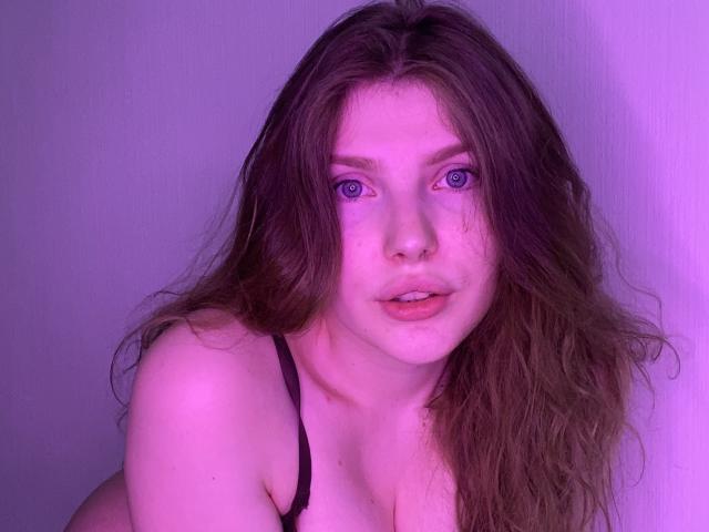 Aleksa Keller ha lunghi capelli castano ondulati, occhi azzurri e un piercing al naso. Indossa un reggiseno nero, con il seno ben visibile, e una luce viola proietta un bagliore soffuso..
