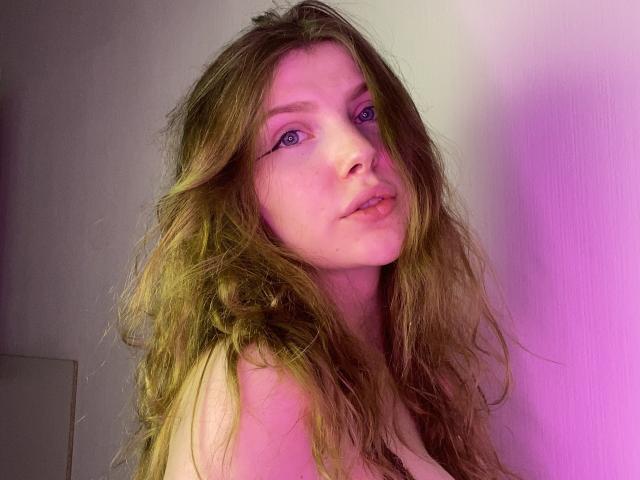 AleksaKeller has long, wavy brown hair, pink eyeshadow, and a slightly open mouth with a small piercing on her bottom lip. She's looking to the left with a pink glow.