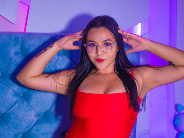 AnaSimpsons with long black hair, red tank top, glasses, and red lipstick, arms raised, hands on head, showing tattoo on right forearm, sitting on blue velvet headboard.