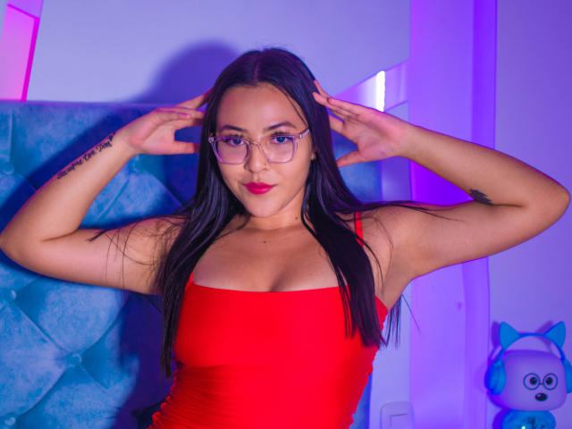 Ana Simpsons avec de longs cheveux noirs, une robe rouge, des lunettes et du rouge à lèvres rouge, les mains sur la tête, un tatouage sur le bras gauche, assise sur un fauteuil bleu capitonné, avec des lumières violettes et roses..