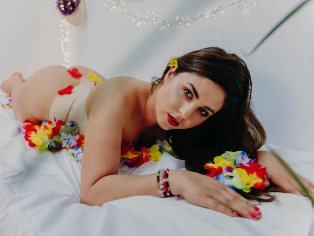 BelleInconnu, lying on white bed, wearing beige panties, surrounded by colorful flowers, looking at camera with red lipstick, brown hair, yellow flower in hair, and red nail polish.