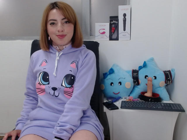 EmillyB con i capelli castano corti, felpa viola con cappuccio a forma di gatto, seduta su una sedia nera, sorridente, gatti di peluche blu con un oggetto simile a un dildo sulla scrivania, parete bianca e una tastiera sullo sfondo..