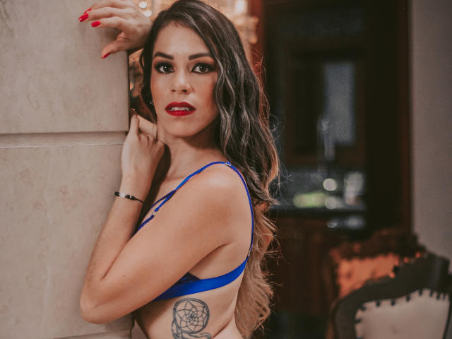 MiaKambell con lunghi capelli castani ondulati, rossetto rosso, reggiseno blu, tatuaggio sul fianco, unghie rosse, appoggiata al muro, che guarda la fotocamera, sexy, sicura di sé, elegante..