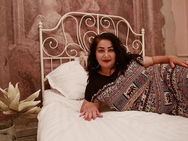 LexyRose with curly dark hair, lying on a white bed, wearing a patterned dress, black top, and hoop earrings, showing her ass and smiling at the camera.