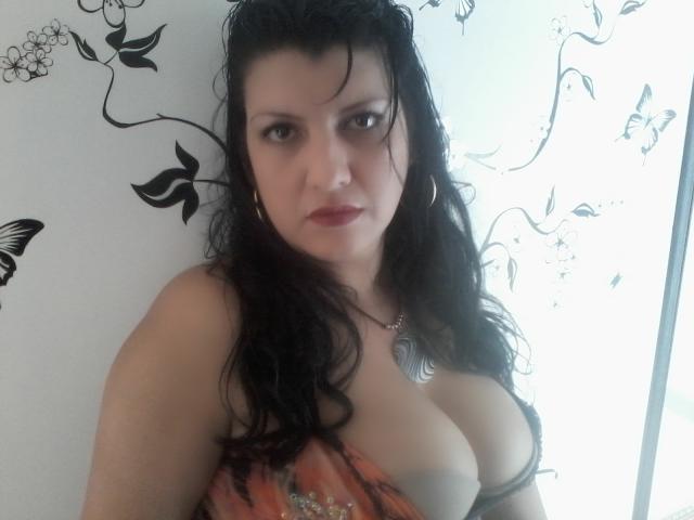 KaterinM, avec ses cheveux noirs mouillés, regarde l'appareil photo. Elle a de grandes seins proéminents, porte une robe orange à décolleté bas, une chaîne en argent et a une expression sérieuse..