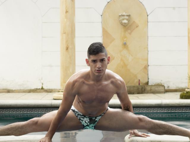 Jonathan Jeremiah, torse nu, musclé, peau bronzée, cheveux bruns courts, assis en tailleur au bord de la piscine, portant un maillot de bain moulant noir et blanc, regardant la caméra, ligne d'eau sur les cuisses, tatouage sur l'avant-bras gauche..