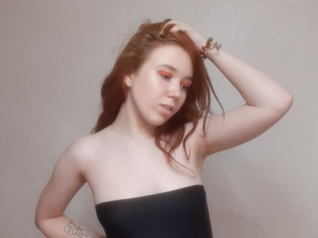 AnikaGlow with red makeup, pale skin, and brown hair, hand in hair, wearing a black strapless top, showing a tattoo on her arm, looking down, soft light.