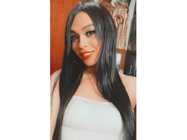 SexyHannaD with long black hair, red lipstick, hoop earrings, and a white strapless top, smiling at the camera. Boobs are visible but not the focus.