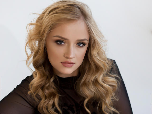 OhMollyHoney with blonde wavy hair, wearing a sheer black top, has blue eyes, full pink lips, and a serious expression. Her tits are small and her ass is not visible.