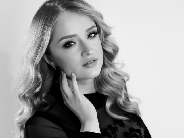 OhMollyHoney has wavy blonde hair, wearing a sheer black top, touching her chin with a delicate hand, in a black-and-white photo with a soft, confident expression.