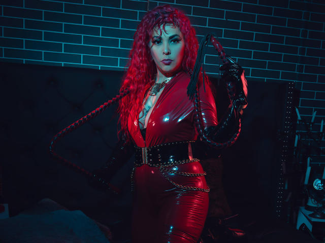 CatheKinkySwitch mit roten Locken, rotem Latekleid, schwarzen Handschuhen, die eine schwarze Lederpeitsche hält, mit Ketten um die Hüfte, vor einem Hintergrund aus einer Ziegelwand, dunkel, stimmungsvoll, sexy, kurvig, mit großen Brüsten..