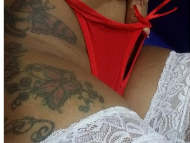A tanga vermelha de KathHott está puxada para o lado, revelando sua vulva tatuada, com meias rendadas brancas que vão até a coxa. Sua mão segura a tanga..
