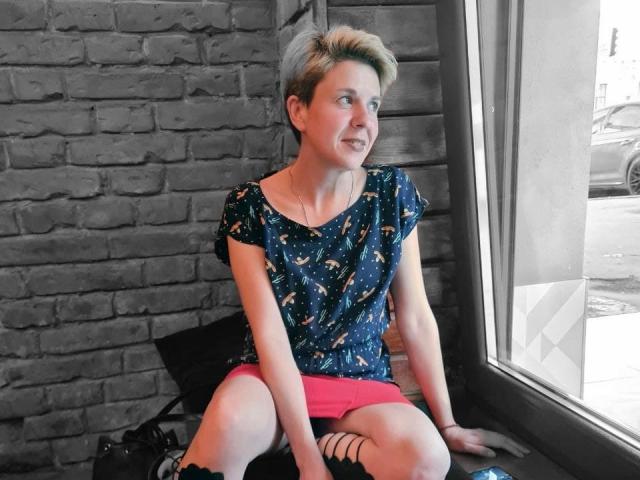 SherlineStone, een vrouw met kort, blond en grijs haar in een pixie-stijl, draagt een blauw shirt met sterrenprint, roze shorts en zwarte sokken. Ze zit op een vensterbank, kijkt naar buiten, glimlacht, met een bakstenen muur op de achtergrond..