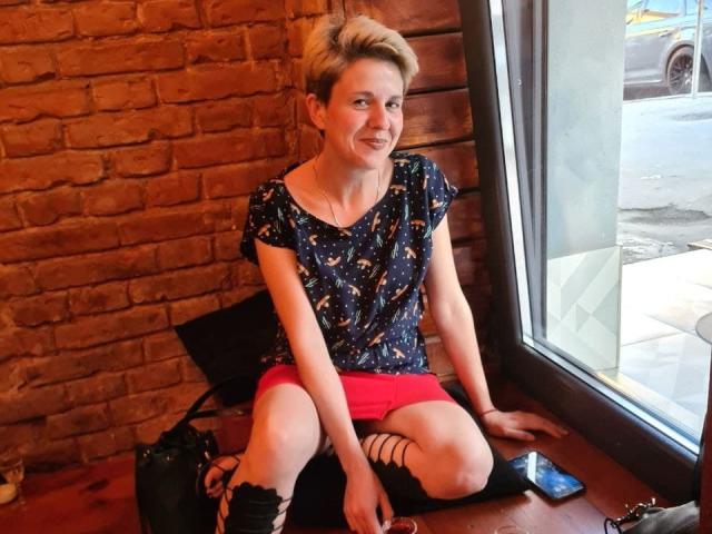 Sherline Stone está sentada en el alféizar de una ventana, vistiendo una blusa azul marino con estampado floral, pantalones cortos rosas, sandalias negras con tiras y un bolso negro. Tiene el cabello rubio corto y una sonrisa..