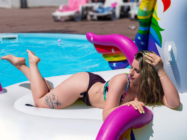 AmyPure is lying on a white inflatable, wearing a colorful bikini, with her ass up and a tattoo on her right thigh. She's holding a pink flamingo.