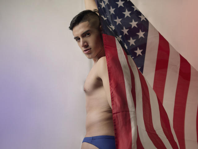JonathanJeremiah, shirtless, wearing blue briefs, holding an American flag, looking at camera, short black hair, light skin, standing, slight smile, flag covering right side of body.