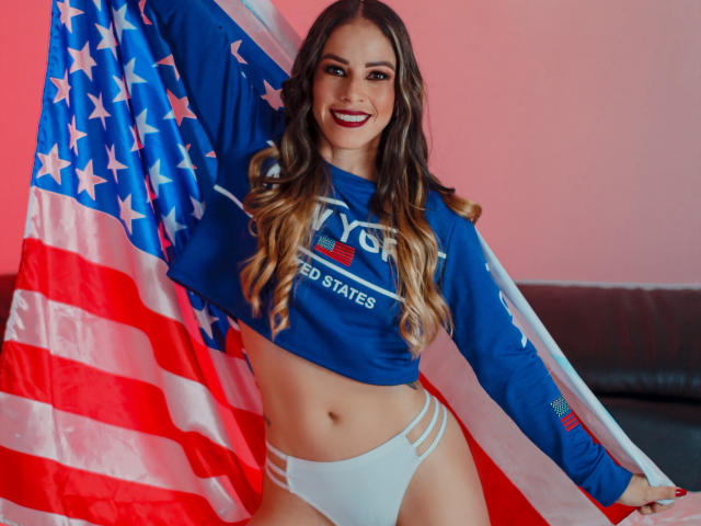 MiaKambell, avec de longs cheveux bruns, du rouge à lèvres, une chemise bleue à manches longues, des sous-vêtements blancs taille haute, tenant un drapeau américain, souriant, montrant ses seins, debout devant un mur rouge..
