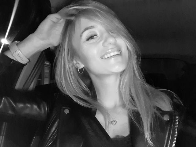 Autoportrait en noir et blanc de LenaaDoll avec de longs cheveux blonds, souriante, portant une veste en cuir, des boucles d'oreilles cercles et un collier en forme de cœur, une main dans les cheveux, regardant vers le haut, avec un anneau au nez..