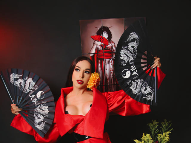 Jhovanna with red kimono, holding black fan with dragon, exposing her big tits, wearing red lipstick, yellow flower in hair, dark background, and red light on left.