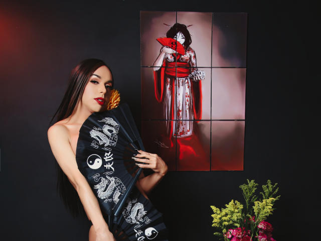 Jhovanna mit langen, dunklen Haaren, rotem Lippenstift, hält einen schwarzen Fächer mit Drachenmotiven und steht neben einem rot-weißen Gemälde einer Frau mit rotem Stoff..
