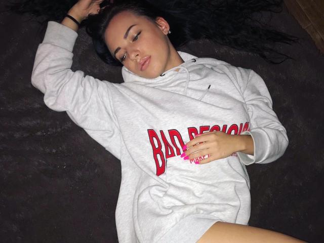 StormTammy ligt op een donker oppervlak, ze draagt een grijze hoodie met rode letters, heeft zwart haar en roze nagels, en heeft haar hand op haar kruis, ze ziet er ontspannen uit..