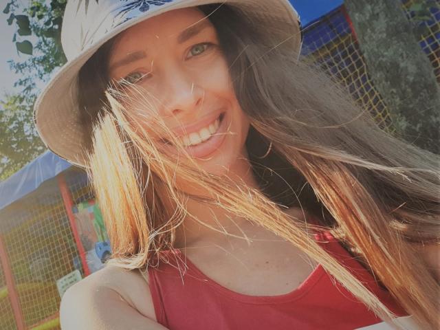MisissFire with long brown hair, smiling, wearing a red tank top and beige bucket hat, sun shining through her hair, green eyes, and cute face, showing her white teeth.