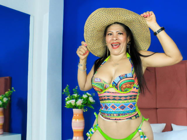 Mara Roberts, com seios grandes, usando um biquíni colorido e geométrico, segurando um chapéu de palha, sorrindo, com uma parede azul ao fundo, alça verde, cama visível, espelho branco e vaso de flores..