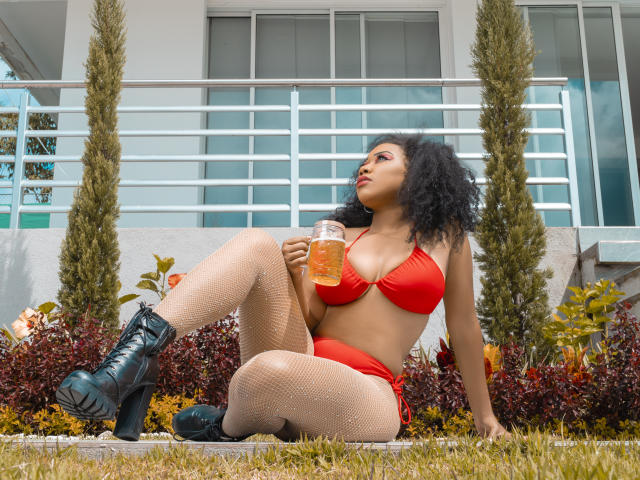 LucyTuker, avec ses cheveux noirs bouclés, son bikini rouge, ses bas en résille et ses bottes noires, tenant une bière, assise sur l'herbe, regardant vers le haut, montrant ses seins et ses fesses..