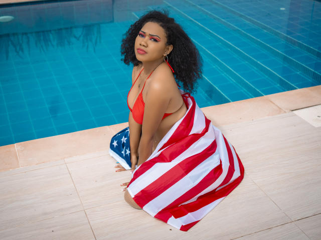 Lucy Tuker, com cabelo preto encaracolado, biquíni vermelho e uma bandeira americana sobre os glúteos, ajoelhada ao lado de uma piscina com azulejos azuis. Ela tem maquiagem vermelha nos olhos e uma expressão confiante..