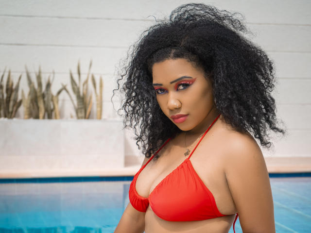 Lucy Tuker mit lockigem, schwarzem Haar, einem roten Bikini und rotem Eyeliner, blickt nach unten und steht an einem blauen Pool, wobei ihre kleinen Brüste sichtbar sind. Ihre Haut ist hellbraun und sie ist süß..