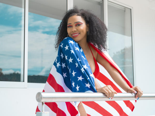 Lucy Tuker, con capelli neri ricci, indossa un bikini rosso, tiene la bandiera americana, sorride, è in piedi su un balcone, con un muro bianco e una finestra sullo sfondo, ha l'ombretto rosso e le unghie blu..