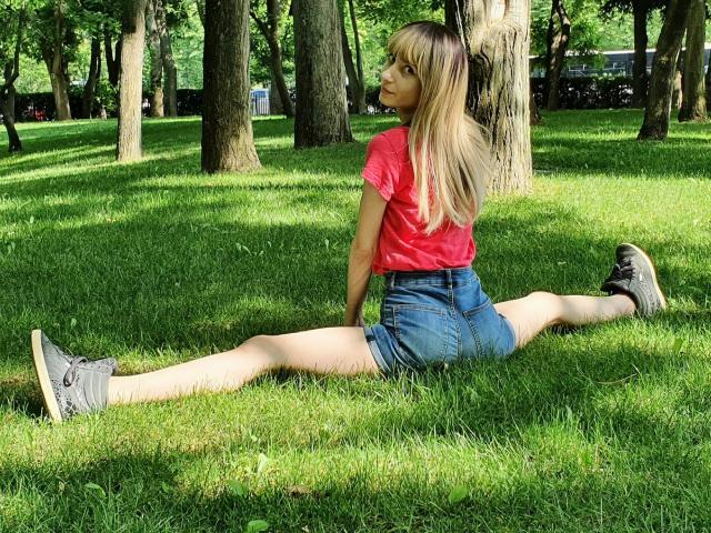 GinnyGold, bionda, con una maglietta rosa, pantaloncini blu e scarpe da ginnastica grigie, sta facendo una spaccata completa sull'erba, mostrando il sedere e le gambe lunghe, in un parco soleggiato, guardando oltre la spalla..