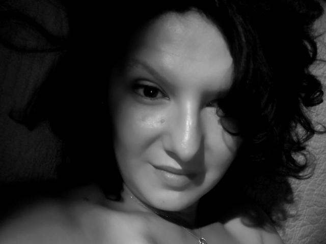 Black-and-white photo of AmethystCharm with curly dark hair, light skin, and a subtle smile, showing her bare shoulders and a necklace, looking directly at the camera.
