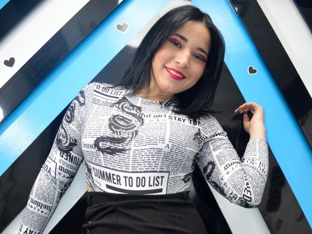 VanessaDubois met zwart haar, rode lippenstift en zwarte eyeliner, draagt een top met een wit en zwart patroon, glimlachend, staand voor blauwe en zwarte geometrische vormen, met haar rechterhand omhoog..