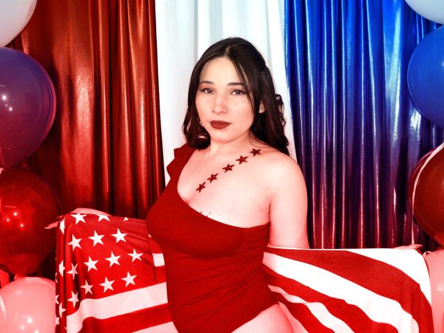 VanessaDubois with dark hair, red lipstick, and red one-shoulder dress, stars on shoulder, American flag in hand, surrounded by red, white, and blue balloons. Boobs visible.