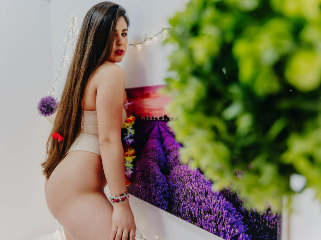 BelleInconnu with long brown hair, wearing a beige thong, showing her ass, looking back with red lipstick, green bracelet, and colorful floral accessory, next to a purple flower wall.