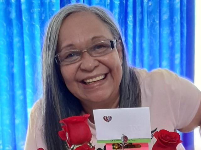 Rosario Triana, com cabelos grisalhos, óculos e um sorriso, segurando rosas vermelhas e um cartão branco com um logotipo de coração, vestindo uma camisa rosa, em um fundo de cortina azul claro..