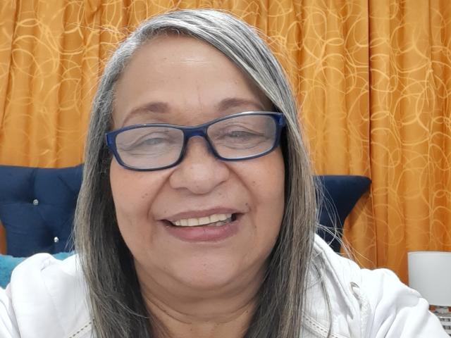 Rosario Triana, con cabello gris, gafas azules y una sonrisa, vestida con una camisa blanca, tiene un rostro regordete y está sentada frente a cortinas amarillas..