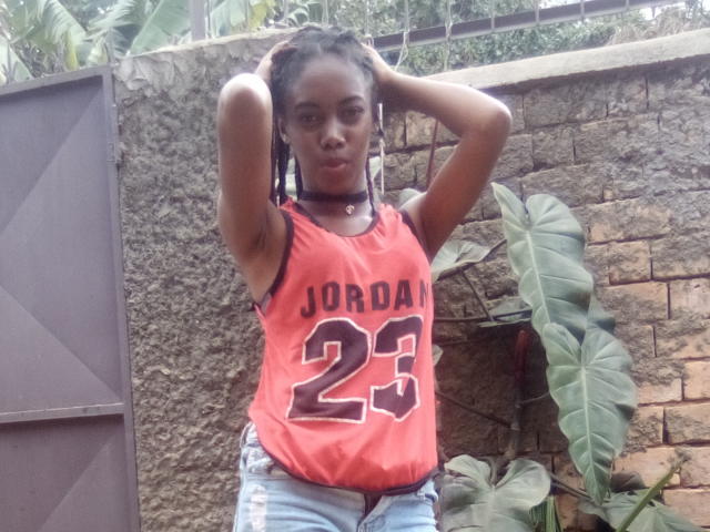 Santamince has dark skin, small tits, and wears a red Jordan 23 jersey with blue jeans, standing with hands in braids, looking at camera, in a backyard.