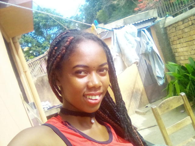 Santamince with dark brown skin and red-black braided hair, smiling, wearing a red tank top and black choker, showing shiny eyes and small nose. Her tits are small.
