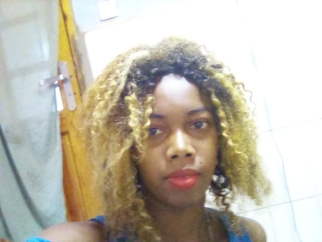 Santamince has dark brown skin, curly blonde and black hair, red lipstick, and is wearing a blue top. She's looking slightly to the side with a neutral expression.